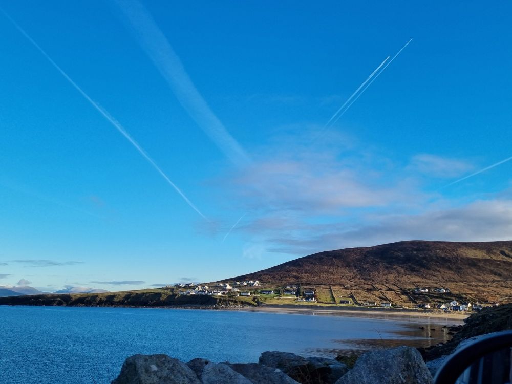 Landscape image taken near the pier in Dugort, Achill Island. The angle faces from the pier towards the village and beach. The sky is mostly clear and very blue. There are three faint lines in the sky that show planes flew overhead shortly before the picture was taken. It's very sunny but it was a crisp, cold day
