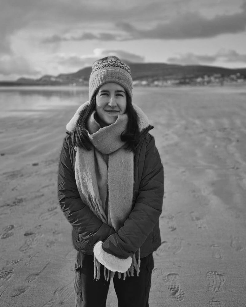 Grayscale image taken of me on Keel Beach, Achill Island, in portrait format. The background is blurred. I am short in stature with shoulder length dark hair. I am wrapped up in a warm coat, scarf, and hat. 