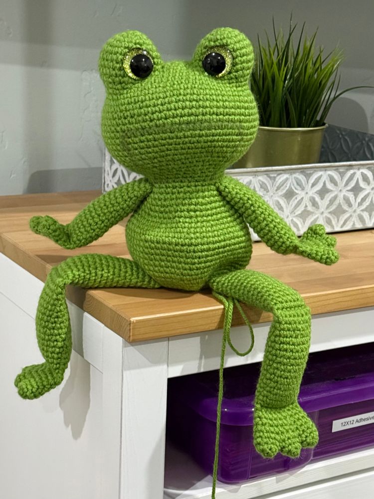 A green crocheted frog with large light green sparkly safety eyes sitting on the corner of a tabletop. The frog has long jointed legs and they dangle off the edge of the table.