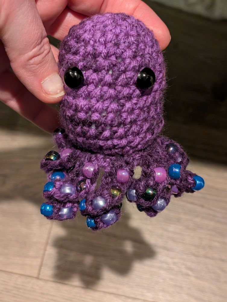 A hand holds a small, crocheted purple octopus with large black eyes against a backdrop of a dark brown wooden floor. The octopus has eight tentacles, each adorned with alternating blue and pearlescent purple beads.  The tentacles are curled and positioned in various directions. The hand holding the octopus is fair-skinned with short, unpolished fingernails. The octopus casts a small shadow on the floor.