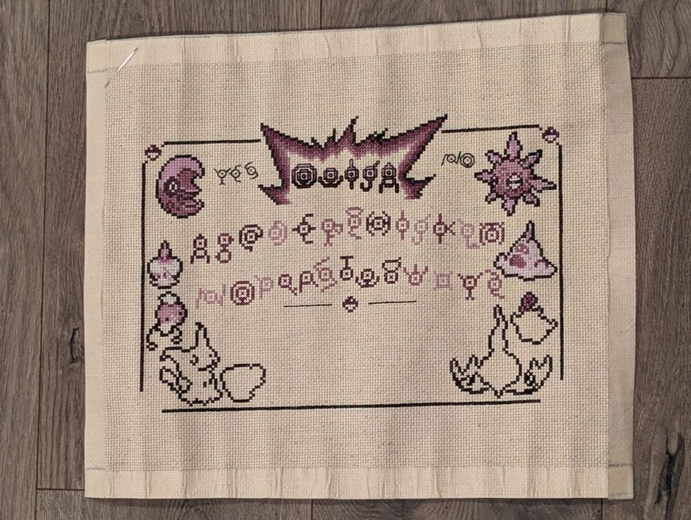 A cross-stitch in shades of purple and black thread depicts a Pokémon-themed Ouija board.  Instead of the usual letters, the alphabet is formed using Unown, with various Ghost-type Pokémon surrounding the board.