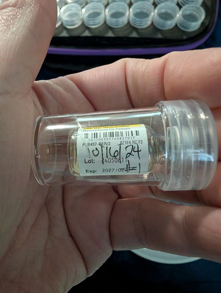A close-up photo shows a hand holding a small, clear plastic jar with a clear lid. The jar contains an empty glass vial, its label partially visible, indicating it once held testosterone cypionate injection. The label shows a manufacture date, with "10/16/24" handwritten next to it, and an expiration date of February 2027.  A handwritten "#1" on the label marks this as the first vial of testosterone used.