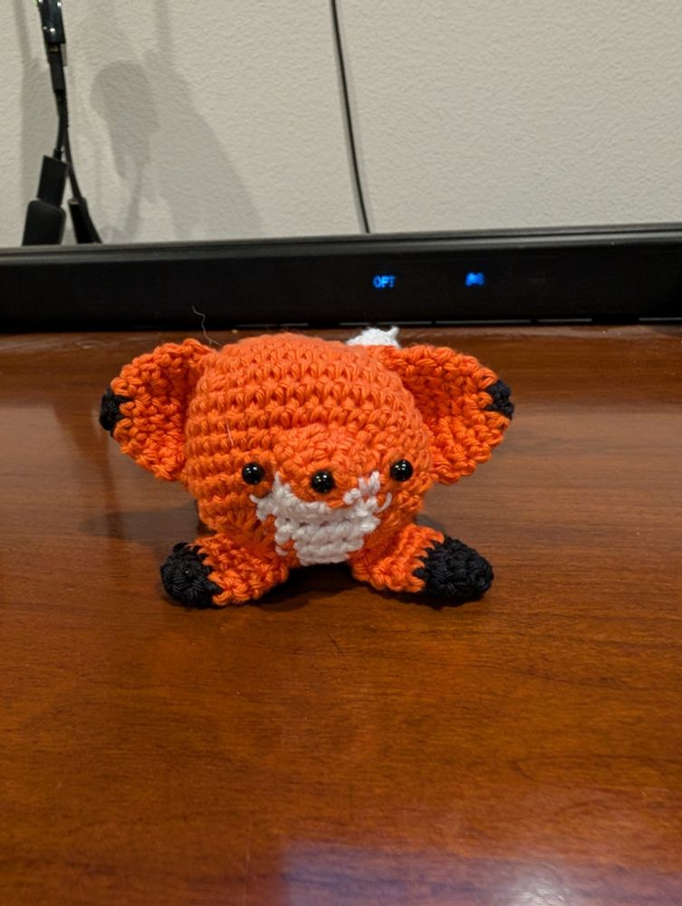 A small, adorable crocheted fox sits straight on on a dark brown wooden table. The fox, crafted using single crochet stitches, is made with orange yarn, with black yarn for its feet and facial features. It has a white snout and small, black bead eyes. The fox is facing the camera in a simple, minimalist design.