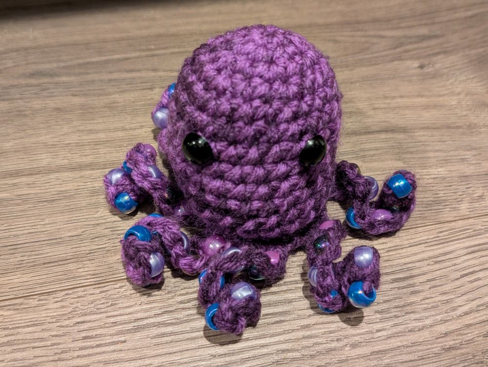 A small, crocheted purple octopus with large black eyes sits on a dark brown wooden surface. It has eight tentacles, each adorned with alternating blue and pearlescent purple beads. The tentacles are curled and positioned in various directions, giving the octopus a playful look. The octopus is positioned slightly angled to the left, and the lighting appears to be coming from above.
