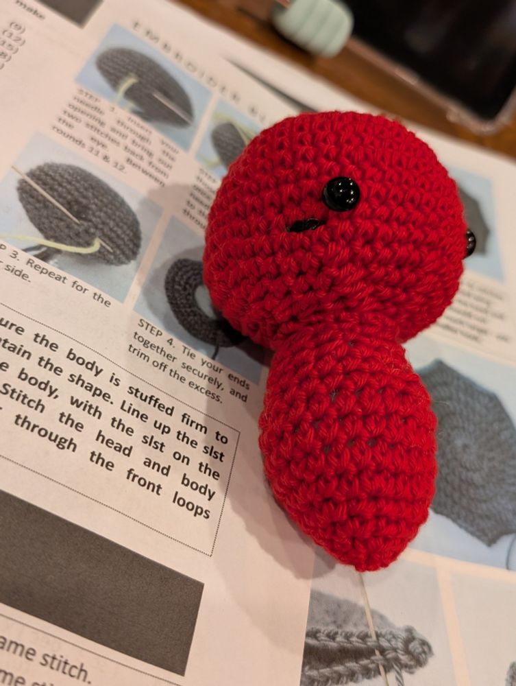 This close-up photo reveals delicate details of a crocheted creation in progress. The focus is on the creature's face, where soft, rosy blush has been delicately embroidered onto the vibrant red yarn.  Two black safety eyes peer out, hinting at the personality that will emerge as this piece is completed. The surrounding pattern instructions, though blurred, show diagrams and text that guide the construction. This intimate perspective, with its soft lighting, invites viewers to appreciate the artistry and anticipation of a handmade work taking shape.