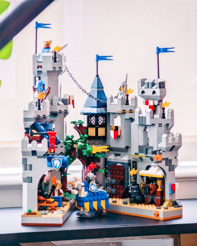 A small folding LEGO castle sits partially folded on a modern shelf in an apartment. The castle has two halves- on the left, a cook prepares a feast under a colorful awning with a residential room above. In the center, a LEGO tree splits open, with one side shadowed by a massive tower. A chain leads from the tower to the other castle half; a pointy tower, a barred wooden door, and a mead stand. A knight on a colorful jousting horse stands guard.