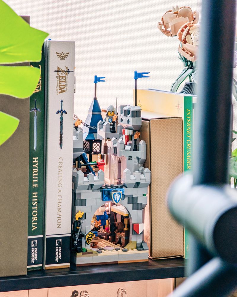 A LEGO castle book nook in closed form; knights peer over battlements and towers in between various books on an a modern apartment shelf.