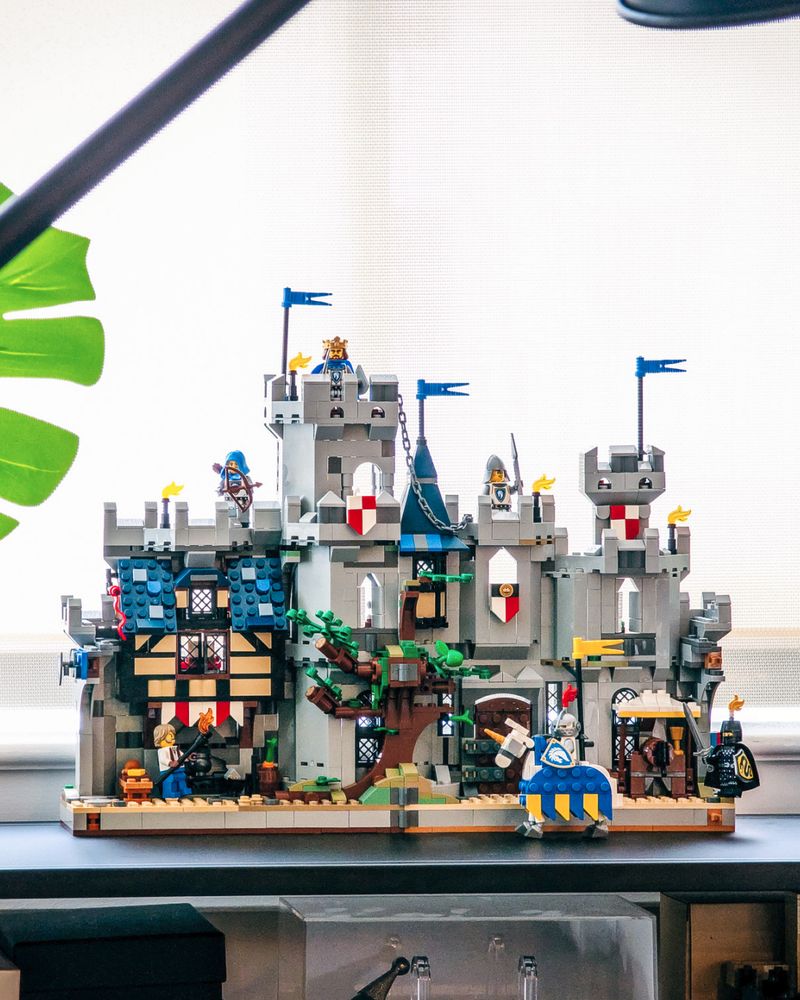 A small folding LEGO castle sits fully open on a modern shelf in an apartment. The castle has two halves- on the left, a cook prepares a feast under a colorful awning with a residential room above. In the center, a LEGO tree splits open, with one side shadowed by a massive tower. A chain leads from the tower to the other castle half; a pointy tower, a barred wooden door, and a mead stand. A knight on a colorful jousting horse stands guard.