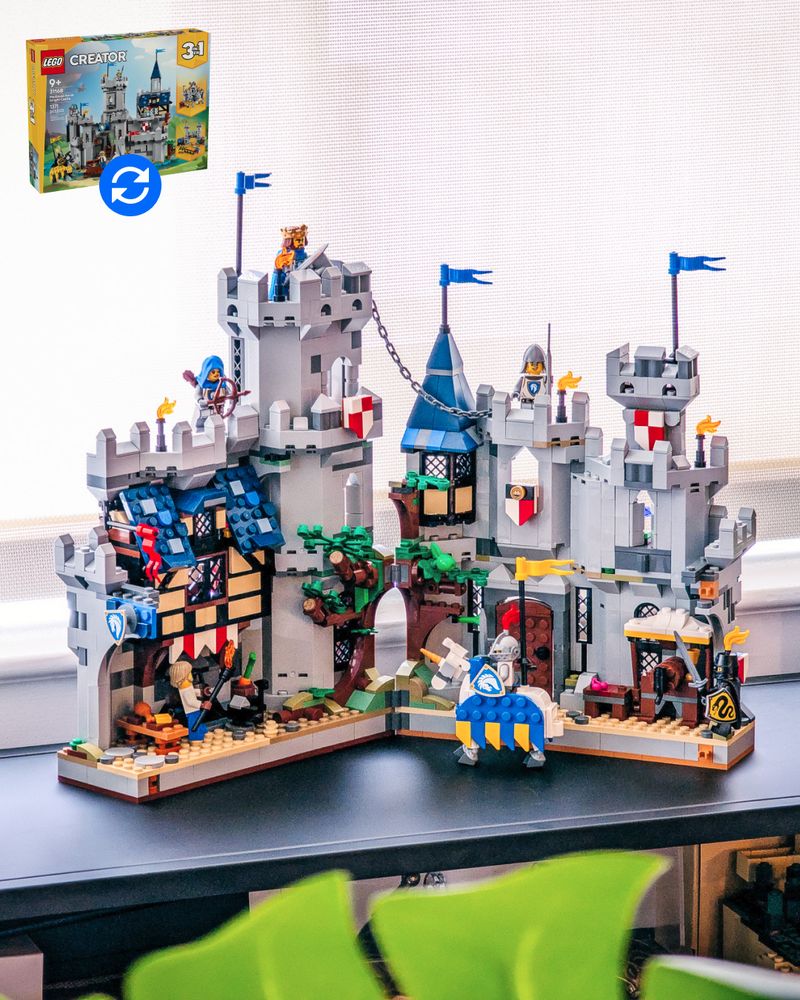 A small folding LEGO castle sits on a modern shelf in an apartment. The castle has two halves- on the left, a cook prepares a feast under a colorful awning with a residential room above. In the center, a LEGO tree splits open, with one side shadowed by a massive tower. A chain leads from the tower to the other castle half; a pointy tower, a barred wooden door, and a mead stand. A knight on a colorful jousting horse stands guard.