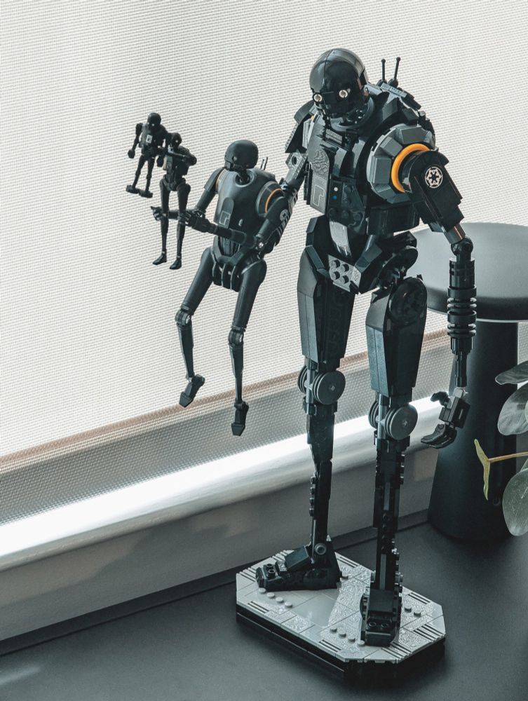A large LEGO-built K-2SO model holds up a smaller Bandai plastic model K-2SO which is in turn holding up a small die-cast TOMY K-2SO which itself is holding up a LEGO K-2SO minifgure, displayed on a metal shelf near a modern lamp and house plant, in an apartment