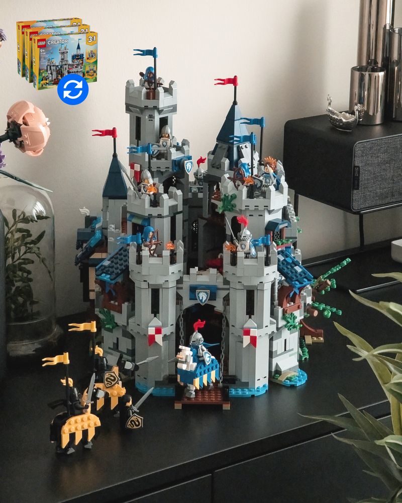 A dense, tall LEGO-built castle sits on a dark dresser inside a modern apartment; the castle features 7 different spires of varying heights; sat within a LEGO-built moat. Three serpent-emblazoned belligerents approach the castle gates, and a horse-mounted knight meets with them pike and shield. Other archers, soldiers, and even royalty observe the melee from the towers above. Artificial plants and black-and-chrome decor surround the castle display.
