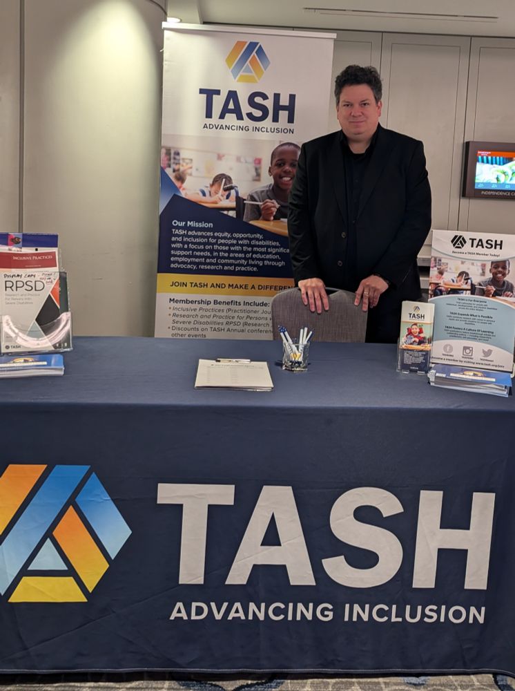 A photograph of a man in a dark suite standing behind an exhibit table covered with a dark blue TASH banner and arrayed with publications and information.