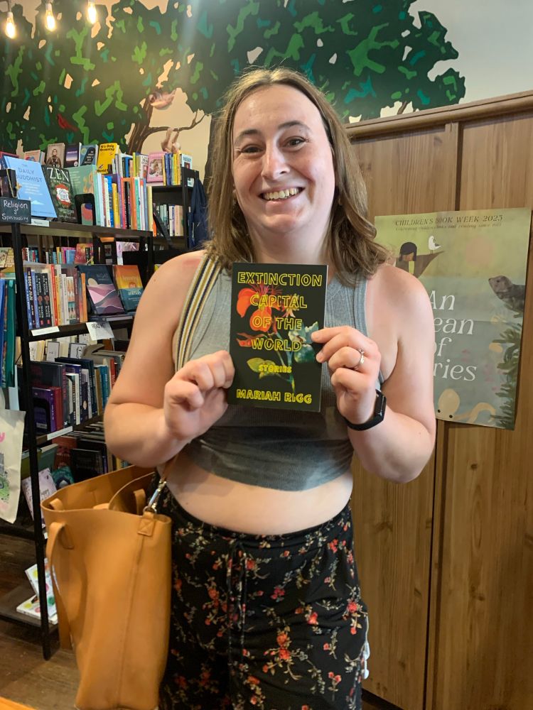 Nic, a brunette white woman, holds a copy of “The Extinction Capital of the World” and smiles 