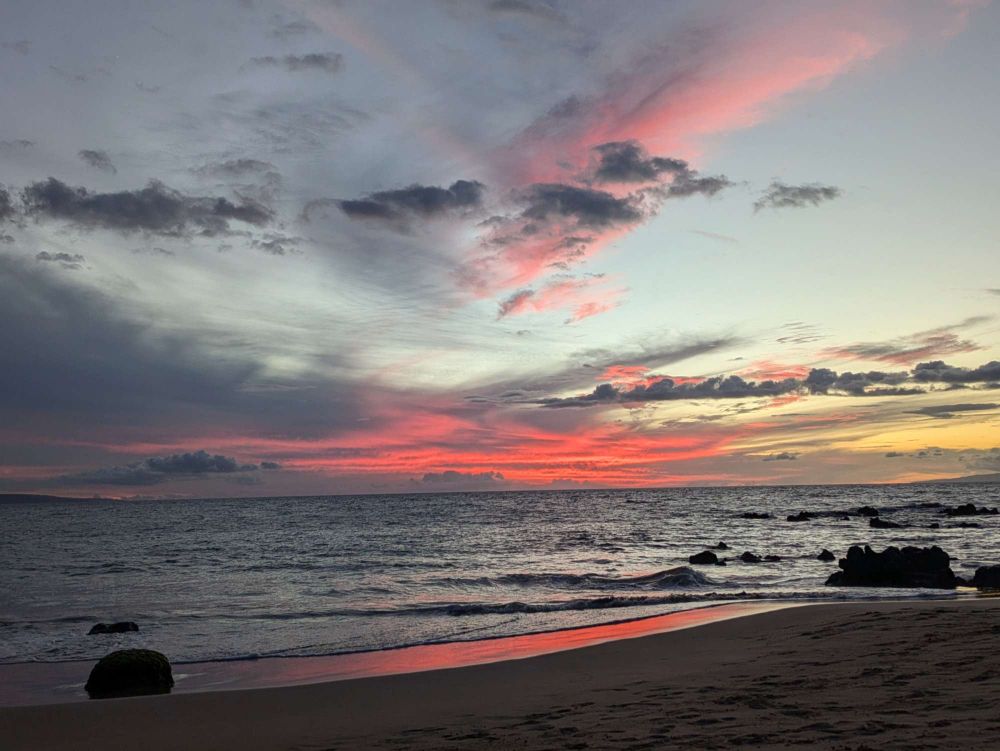 sunset on an ocean beach, deep pink with grey and pink clouds against blue sky. The sunset is reflected at the edge of the water.
