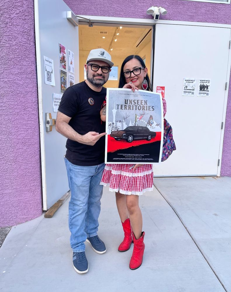 Man with hat and glasses with woman in red dress and glasses posing with a movie poster.