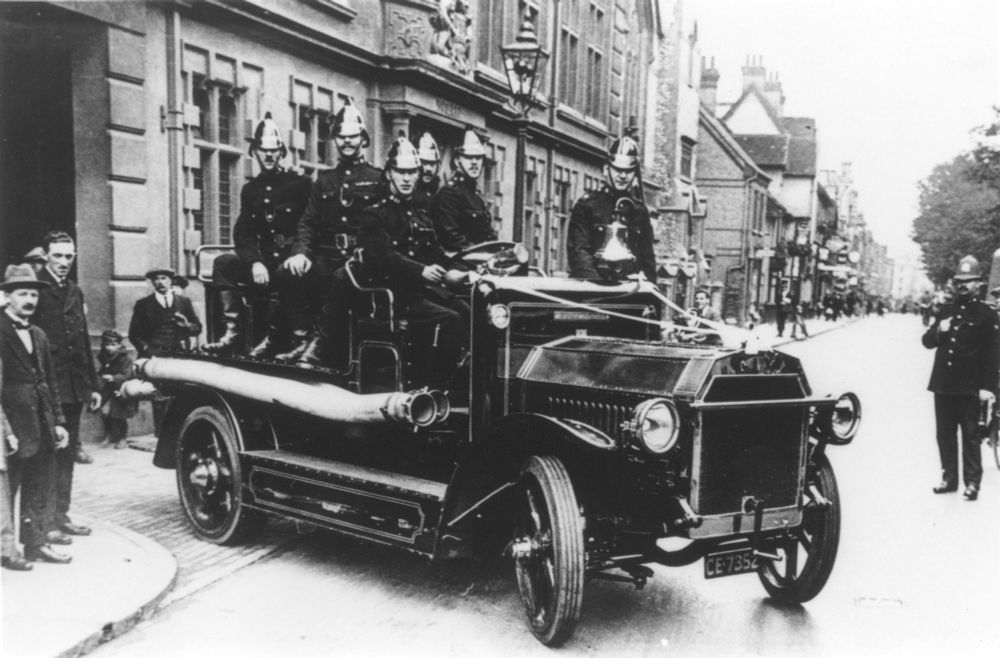 A motorised fire engine in 1920s style leaving the fire station in St Andrew's Street. A full crew of six are perched on top, all looking natty with their shiny helmets.
