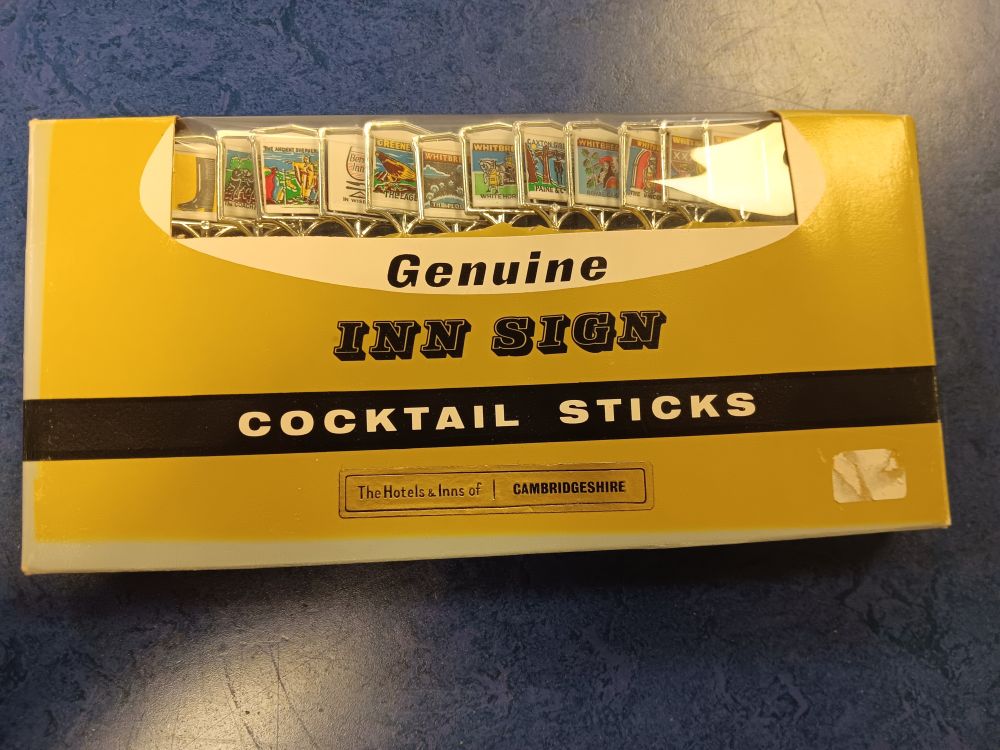 A greeny yellow box, obviously 1960s style, with Genuine Inn Sign Cocktail Sticks The Hotels and Inns of Cambridgeshire from Whitbread. You can see various inn signs sticking out of the top of the box.