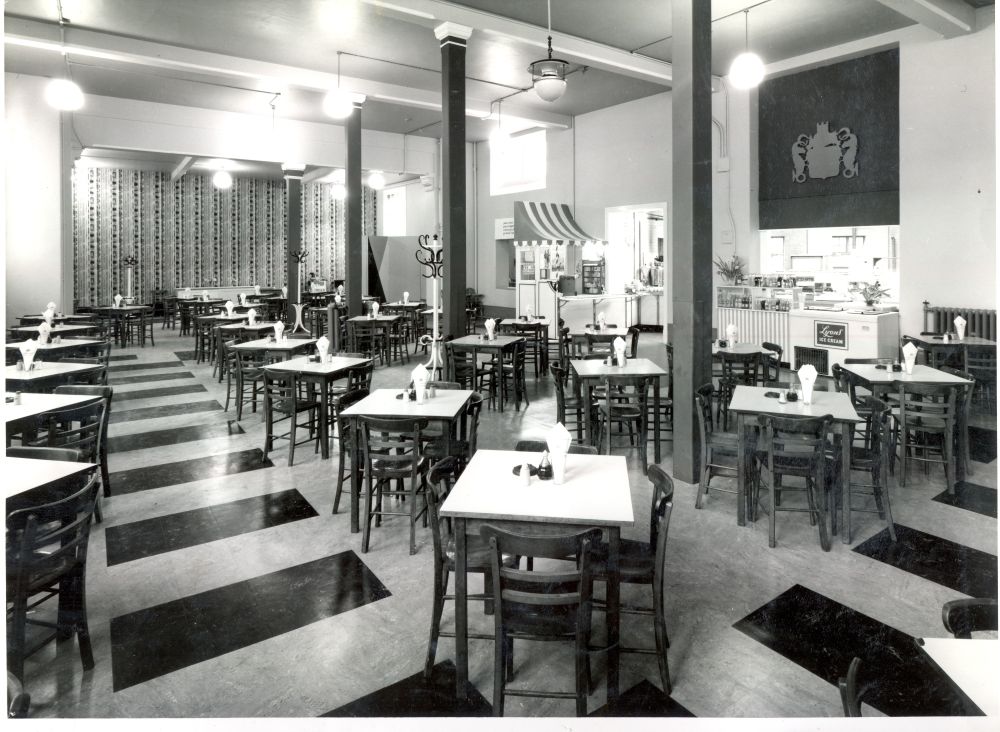 The inside of the Civic Restaurant in 1963. The restaurant is empty, ready to open. There are many square tables laid out in rows, each with four chairs around. The crest of the Cambridge City Council is up on the wall.