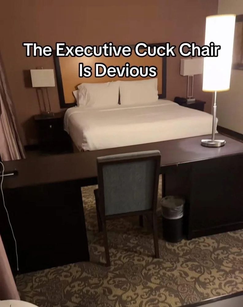 A hotel bed with a desk and chair placed awkwardly at the foot of said bed, slightly off to the left. A meme style caption reads "The Executive Cuck Chair Is Devious."