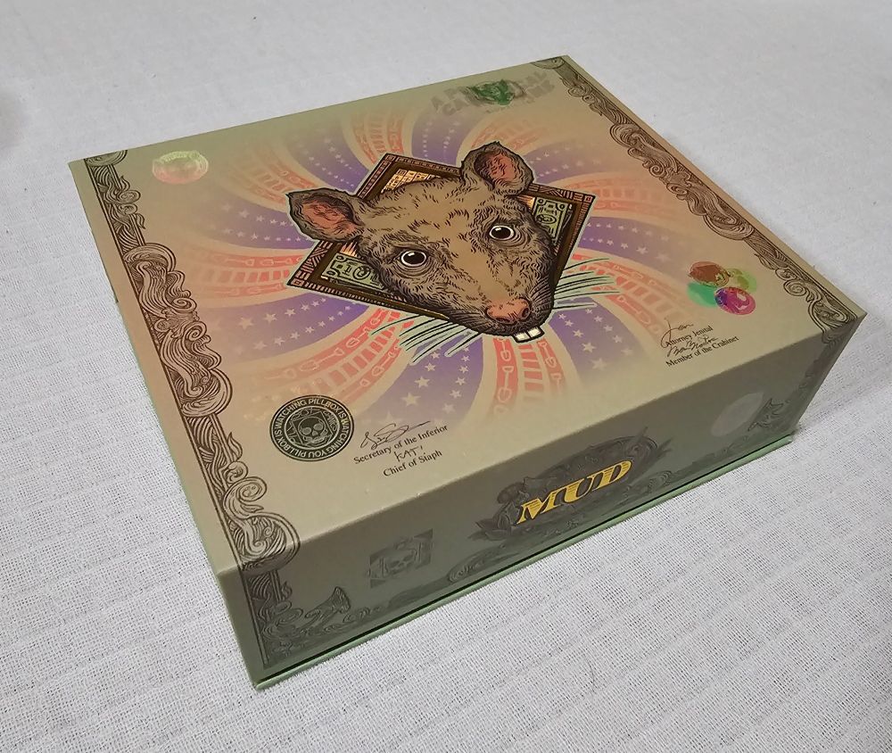 The outer packaging of Pillbox Games' card-based board game Mud, which features an illustration of a rat surrounded by designs pulled from US currency and political posters. The name of the game is emblazoned on the box's fore-edge in gold. The box sits on a white tablecloth. 