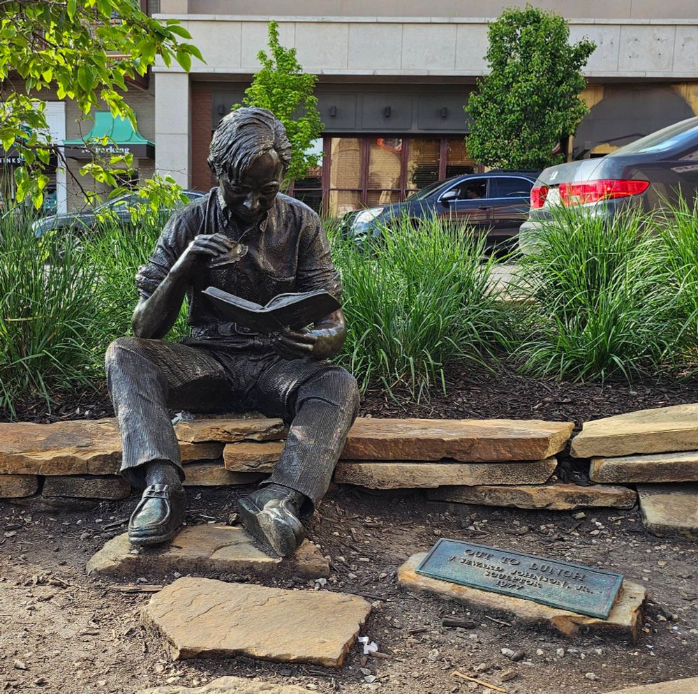 A bronze sculpture of a seated young man wearing chukka boots, corduroy trousers, and a button-up shirt with the sleeves rolled up, his knees splayed thoughtlessly. He holds a hamburger with a bite missing from it in one hand, a rather draggled book in the other, and seems to be reading intently as he eats. The sculpture is seated on a low wall with grassy bushes behind it. In the lower right corner of the photograph, a plate reading "Out to Lunch / J. Seward Johnson Jr. / Sculptor / 1977" can be seen, set atop a flat stone.