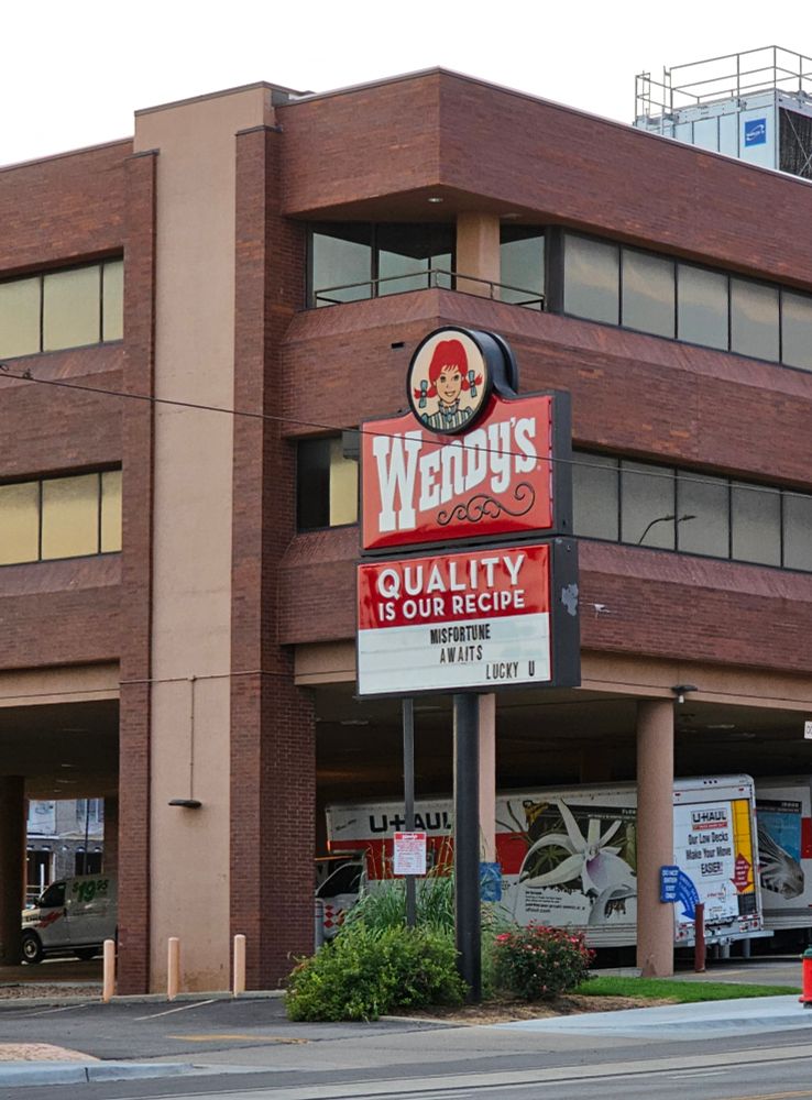 A Wendy's sign, at the corner of an office building with a parking area on the ground floor where a number of Uhaul vehicles are parked. The sign rises from a small grassy area, a few overgrown bushes just behind the supporting pole. The marquee reads:
MISFORTUNE 
  AWAITS
                LUCKY U