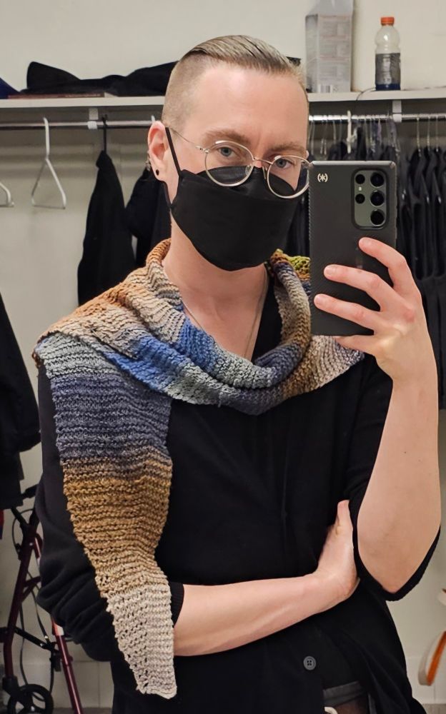 A white person with blonde hair that is buzzed at the sides but long and pulled back on top, dressed in black with exposed forearms, who is wearing glasses, takes a mirror-selfie. They wear a knitted wrap that tapers to a blunt point where it hangs down in front; it has variegated stripes of beige, tan, blues, greens, and browns. The wrap is secured at their left shoulder with a long, silver pin. As evidenced by the dark jackets on hangers, a red-framed walker, and other items in the background, the photo was clearly taken in a workplace bathroom. 
#knitting
#craftsky