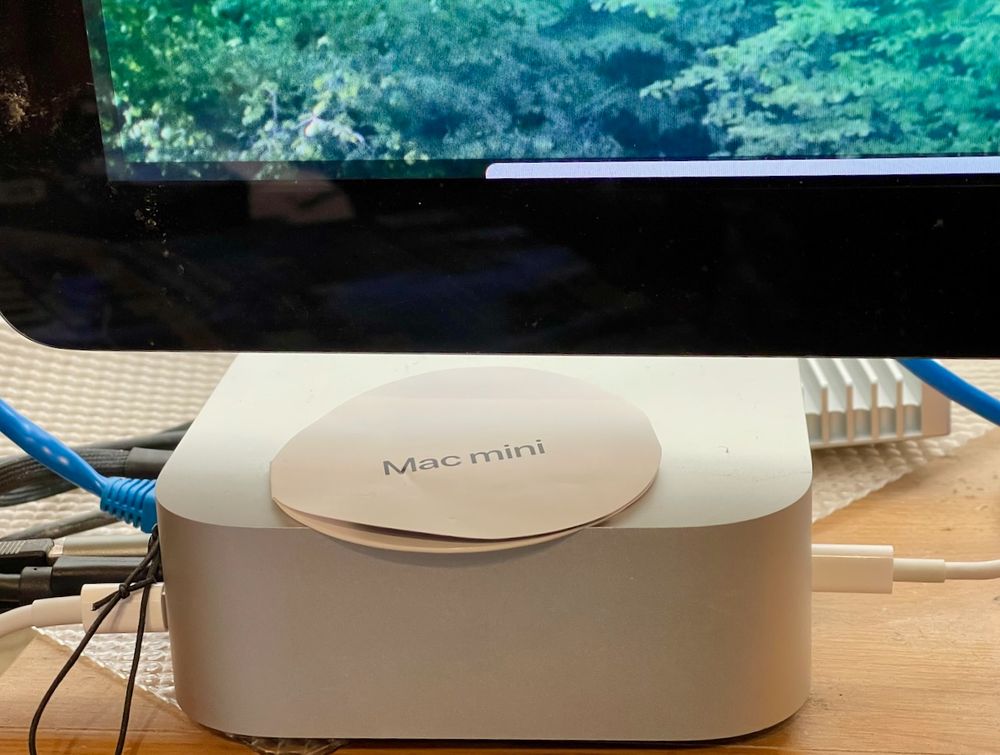 Photo showing an M4 Mac mini connected to various cables and the lower left portion of a monitor displaying the desktop image of MacOS Sequoia.