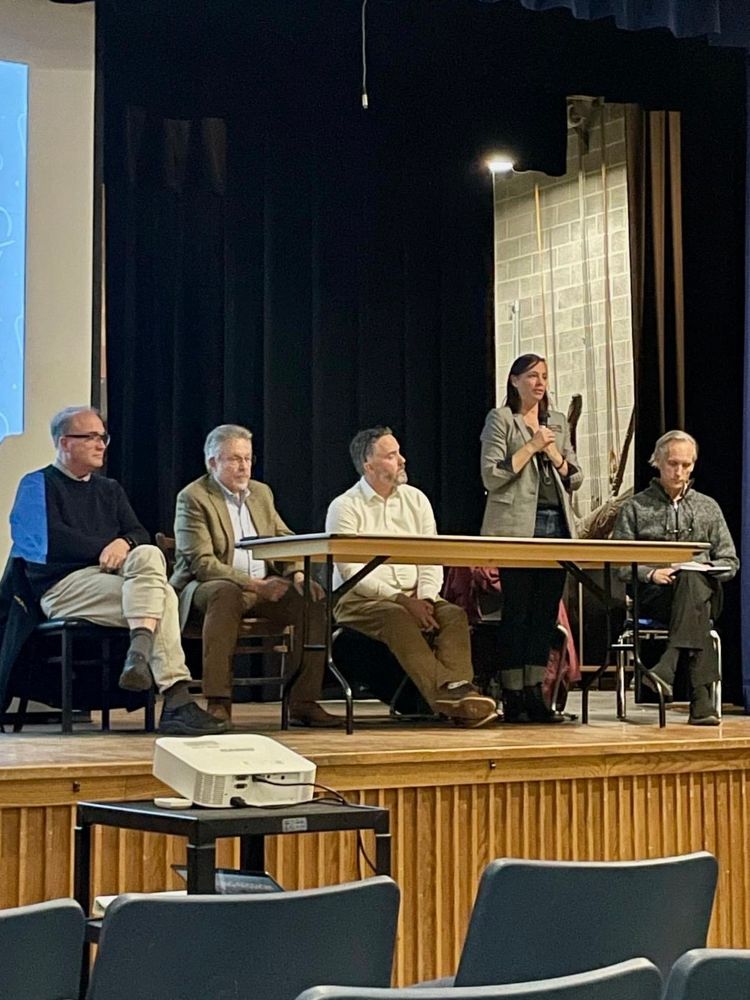 Photo of five leaders sitting at a table elevated on a stage. One woman stands to speak