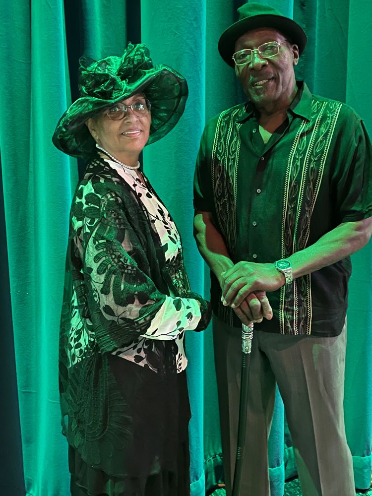 This picture is of Mama Munroe and Papa Munroe. Mama is wearing a big sheer colored hat, white top with black flowers, a black lace shawl, and black skirt. Papa Munroe is wearing a brimmed hat, a patterned button up and slacks. He is also sporting a black cane with a silver top. Both of them are lit up under a green light.
