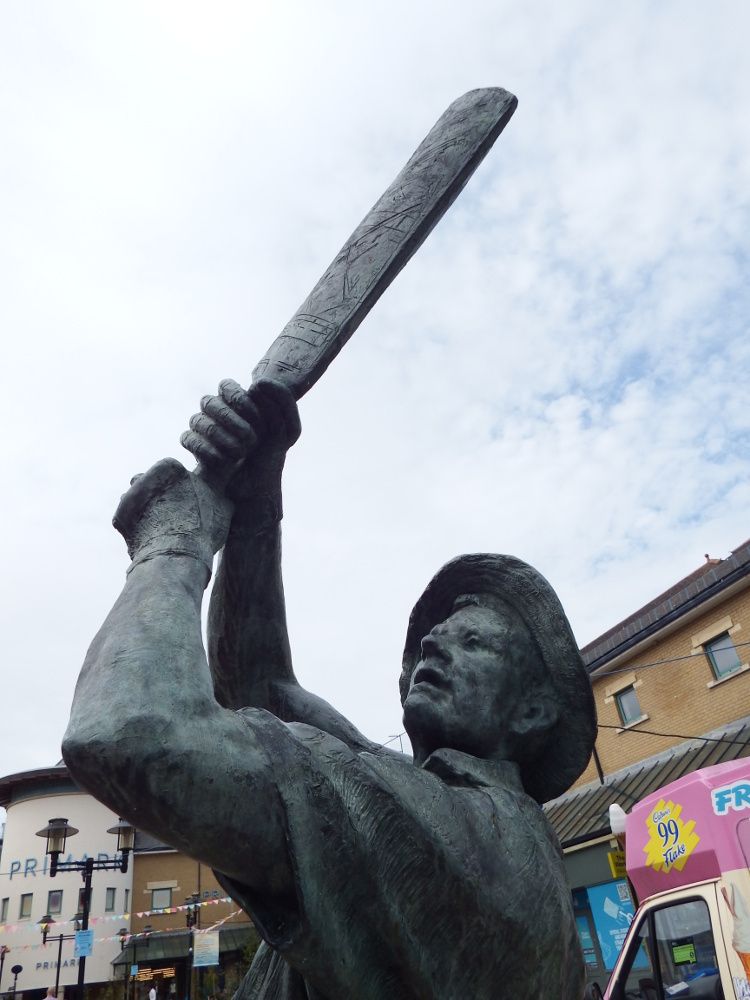 The top of a bronze statue showing a person dressed for cricket holding a cricket bat up, as though just after hitting the ball. An ice cream van and a shopping centre can be seen behind the statue.
