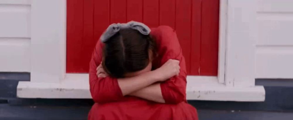 a young girl in a red dress is sitting on the porch of a red building covering her face with her hands .