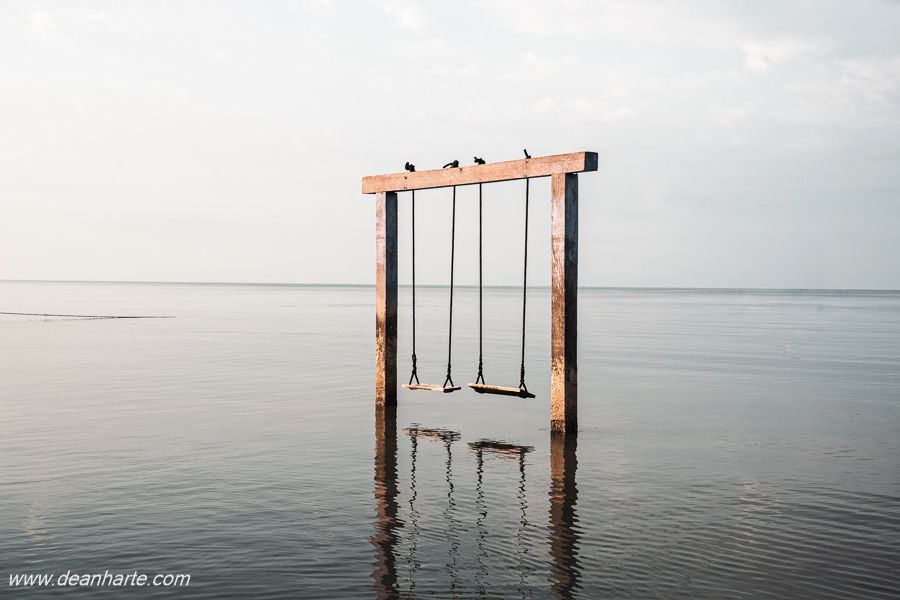 Two empty swings stand above calm, shallow water at low tide in Lombok, Indonesia. The ocean reflects soft light and muted colors, creating a tranquil minimalist seascape that evokes stillness, balance, and tropical serenity.