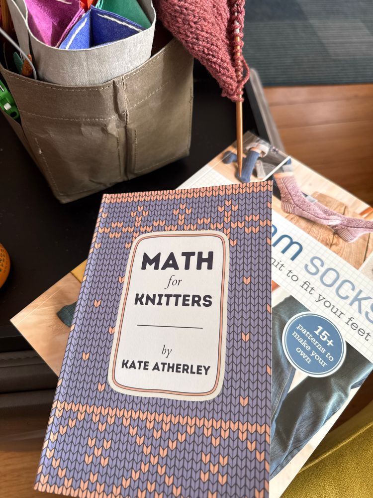 A picture with two books written by Kate Atherley, “Math for Knitters” and “Custom Socks Knit to Fit Your Feet” alongside a small bit of pinkish knitting in a project bag, sitting on an end table.