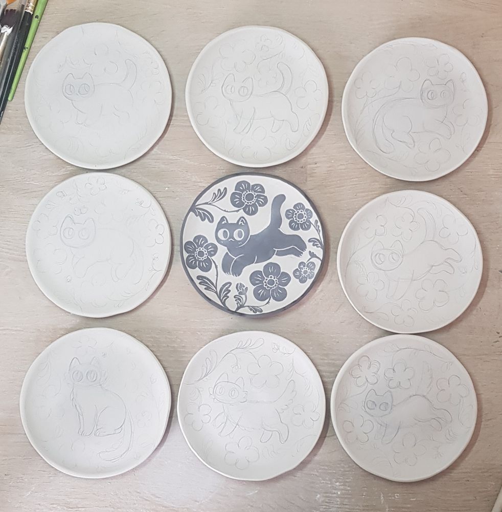 9 unifred ceramic plates, one in the middle is painted with an image of a black cat surrounded by black flowers, the ones around it have pencil sketches on them