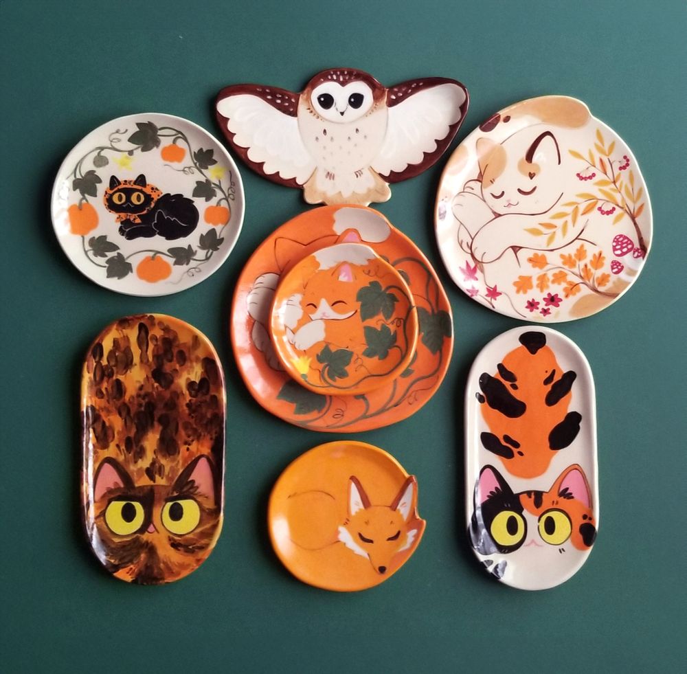 a flat lay with a bunch of ceramic plates of different shapes and sizes, mostly shaped like cats, but there is also a fox and an owl. they're in orange, brown and yellow tones on a dark green background