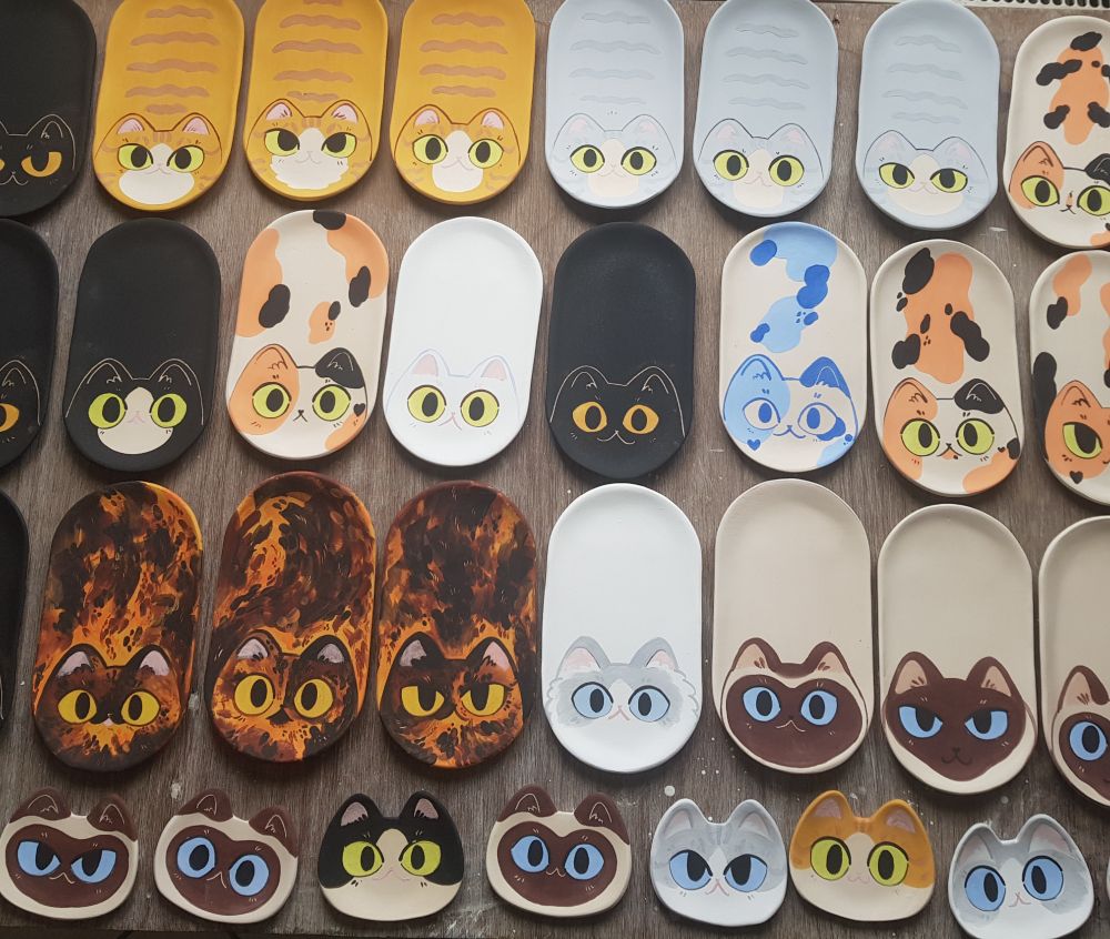 a table top covered with lined up ceramic trays in a shape of loafing cats of various colors