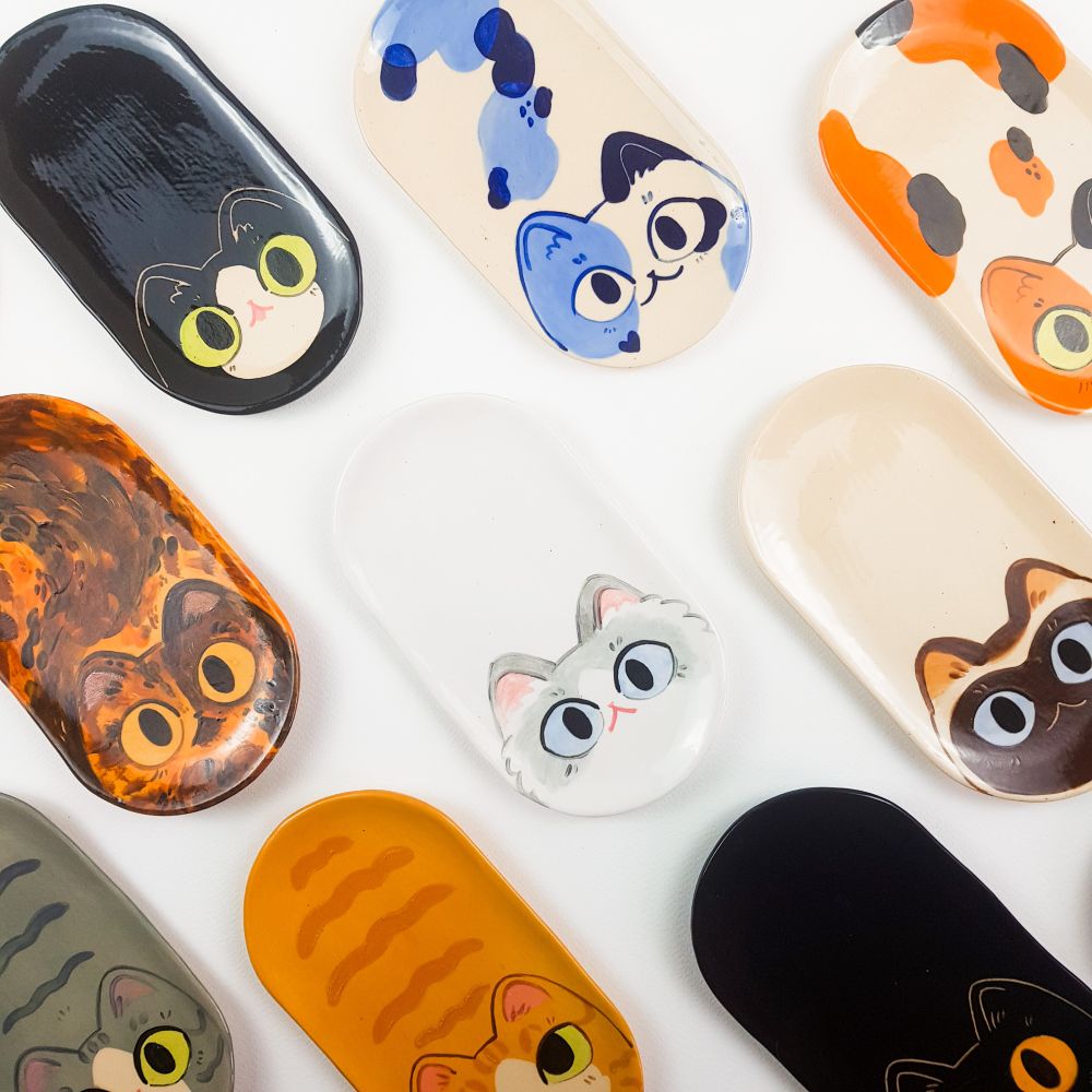 a bunch of ceramic trays shaped like loafing cats lined up on a white background, each cat is different