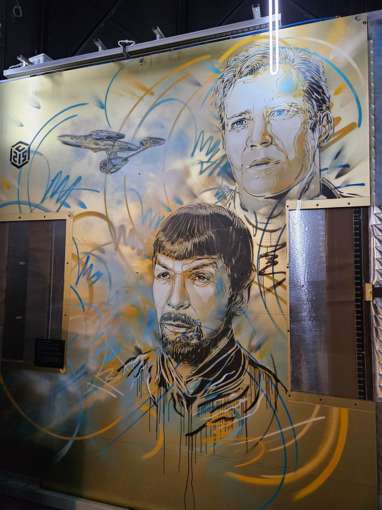 A colorful mural depicting two characters from the Star Trek science fiction series, Spock and Kirk, with the Enterprise flying away in the background. On the left and right of the central figure of Spock, one can notice two wire chambers, particle detectors from the NOMAD high-energy physics experiment that was run at CERN.