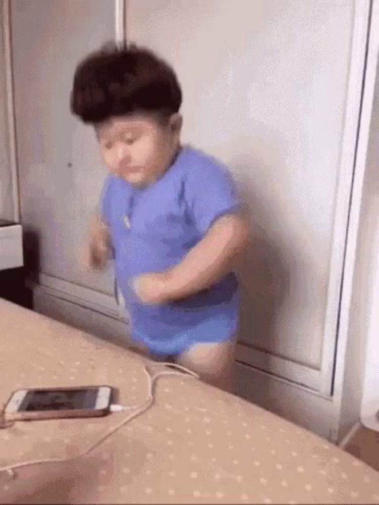 a baby in a blue shirt is dancing on a bed next to a phone .