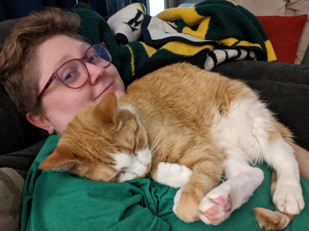 An orange and cream cat is lounging and asleep comfortably on a person with short hair in a green shirt. There are several blankets and pillows in the background. THey are both laying down on a couch.