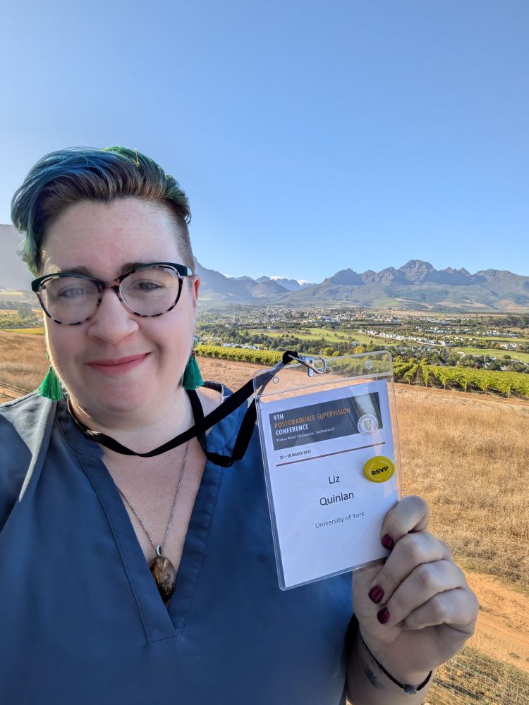 A white woman, Liz Quinlan, stands smiling and holding up a conference badge with her name and an RSVP pin badge on it. In the background are vineyards and a mountain range.