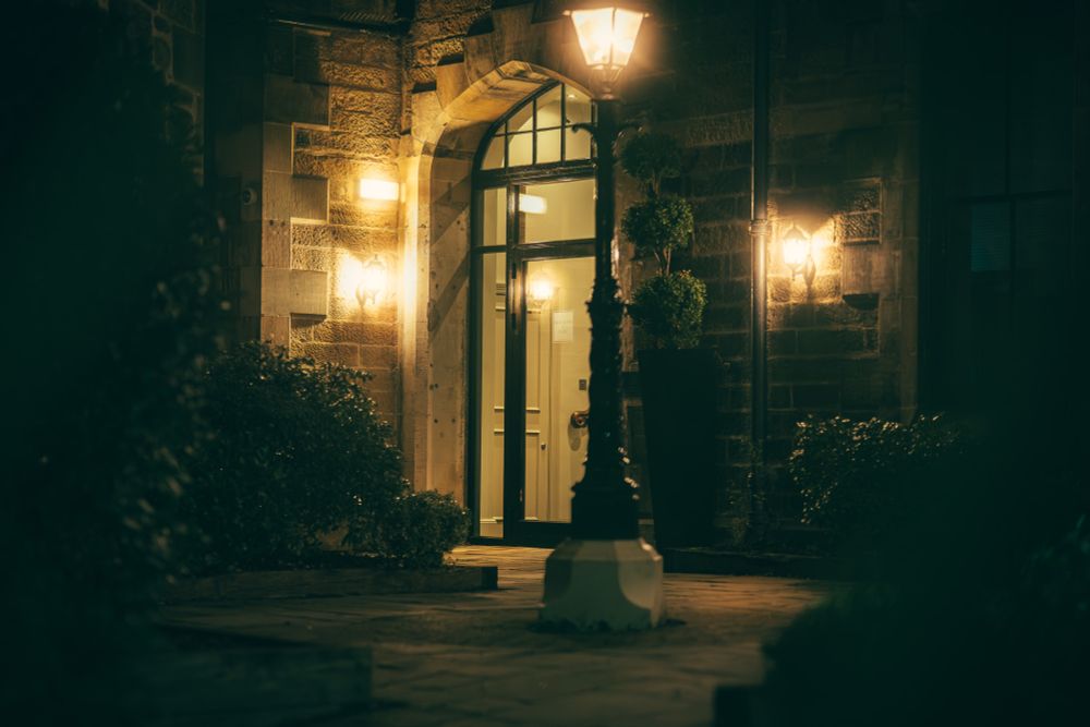 A nighttime view of a stone building entrance with an arched glass door. Two lantern-style wall lights and a decorative streetlamp illuminate the walkway, casting a cozy glow on the stone walls and neatly trimmed bushes.