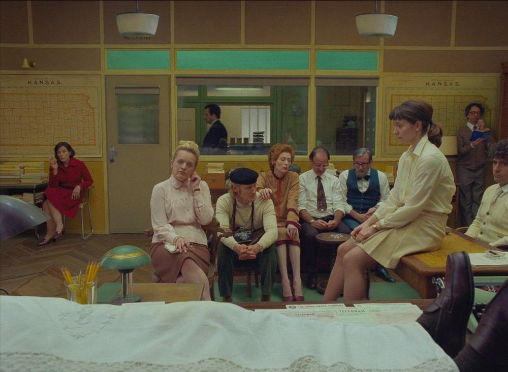 The French Dispatch. The legs of a person covered in a sheet on a desk, many characters sitting on desks and chairs, a man walking by outside the office.