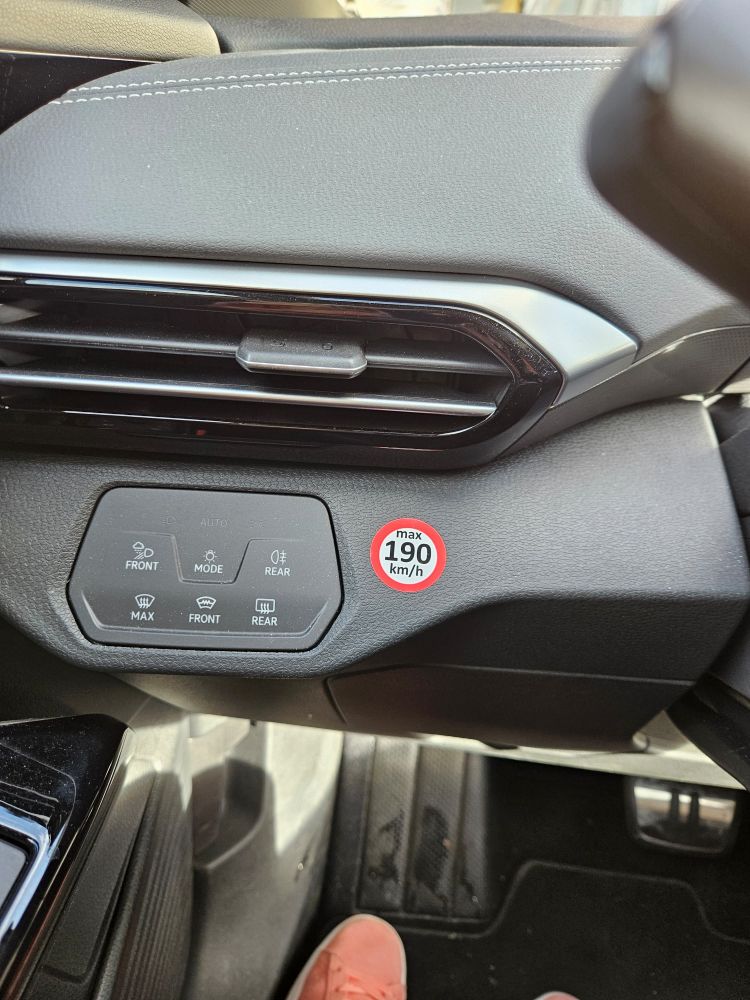 Foto aus dem Auto. Blick aufs Amaturenbrett links vom Lenkrad. Neben den Knöpfen zur Einstellung von Licht und Heizung klebt ein leuchtender runder Aufkleber mit rotem Rand. In der Mitte steht "max 190 km/h".