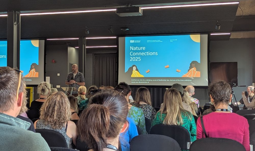Room full of people looking at large screen showing a slide that says Nature Connections 2025. You can see the backs of people's heads and the speaker at the front.