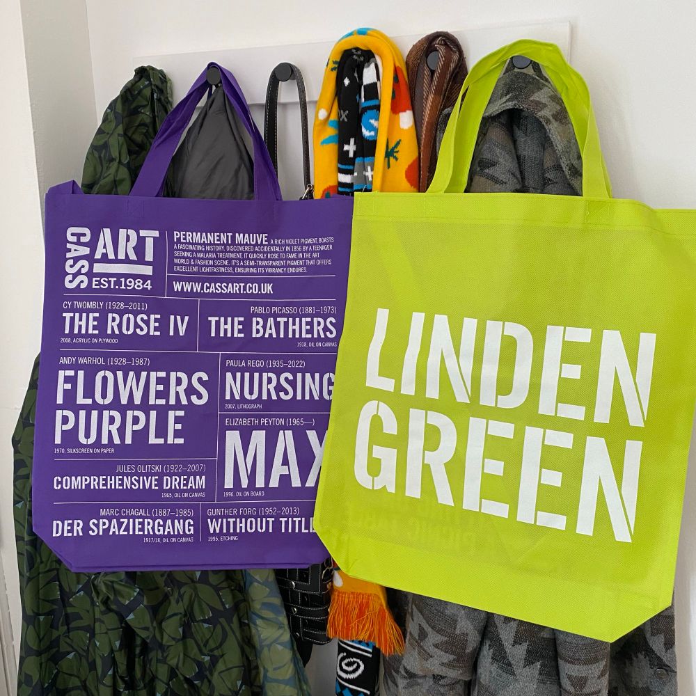 A photograph of a green tote bag and a purple tote bag on a coat hook. They both have lots of graphic text on them 