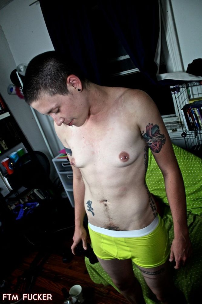 a younger biff is standing shirtless in neon yellow boxer briefs, looking downward. his brown hair is buzzed short, his nipples and navel are pierced. he is skinnier but has definition in his abdominals. the room has hardwood floors and some shelves, and one of the crappiest futon sofas ever made. in the bottom left corner is a watermark for FTM Fucker.