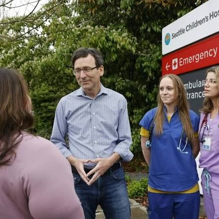 WA state governor Bob Ferguson in blue jeans and button-up shirt standing alongside medical staff in front of Seattle Children's Hospital
