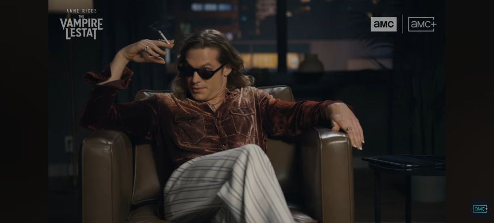O ator Sam Reid caracterizado como vampiro Lestat, usando uma camisa de mangas cumpridas de veludo marrom e calças brancas com listras finas de tom escuro, de corte social. Ele fuma e usa óculos e olha com desdém falando com alguém ao seu lado direito. Ele está sentado em uma poltrona de couro marrom claro, uma mesinha ao seu lado esquerdo, e ao fundo pode-se ver uma janela, mostrando a noite e parcialmente um prédio. Na imagem pode se ver 3 vezes o logotipo "AMC" e "AMC+" que estão ao lado direto da tela, e o outro logo no lado direito, escrito "Anne Rice's The Vampire Lestat"